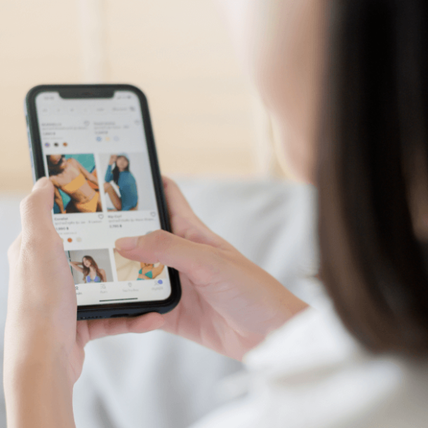 A woman wearing a white shirt holds a smartphone and scrolls through pictures of clothing for sale