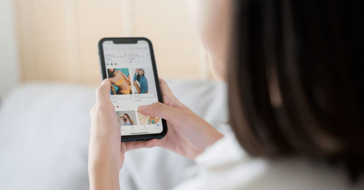 A woman wearing a white shirt holds a smartphone and scrolls through pictures of clothing for sale