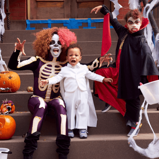 A family of 3 makes scary faces and monster-like gestures while sitting on a cement stoop covered in fake spiderwebs and pumpkins. The mother, a Black woman wears a curly pink and brown wig, white skeleton facepaint and a skeleton cosume. Standing next to her is a young Black child wearing an all-white Dracula costume, and to the right is a boy wearing red hair paint, Dracula face paint, and an equally scary black and red dracula costume. He holds a candy bucket in his left hand and is raising the other in a monster pose above mom and baby brother.