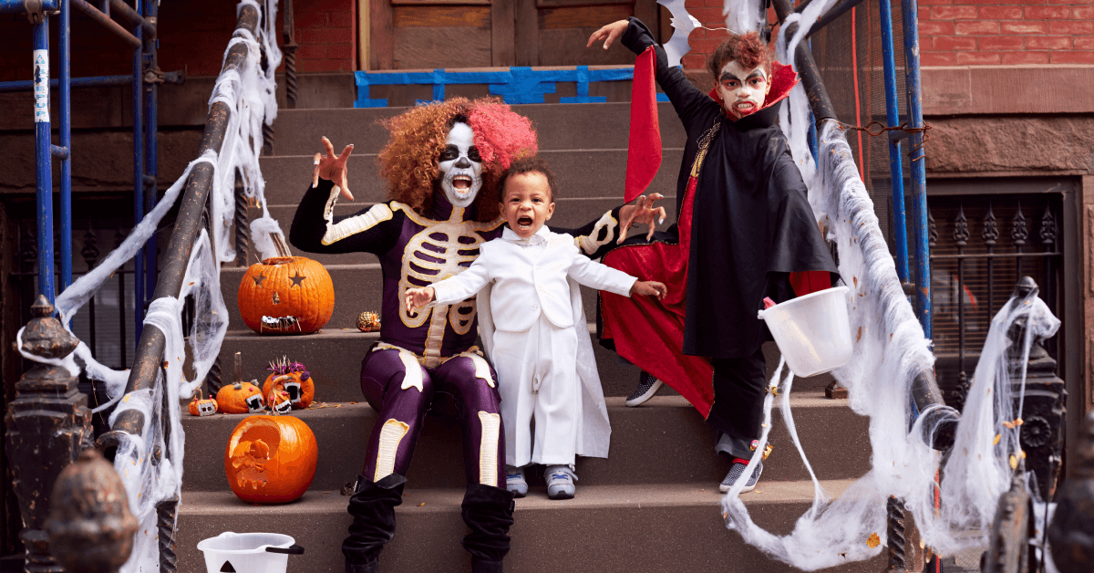 A family of 3 makes scary faces and monster-like gestures while sitting on a cement stoop covered in fake spiderwebs and pumpkins. The mother, a Black woman wears a curly pink and brown wig, white skeleton facepaint and a skeleton cosume. Standing next to her is a young Black child wearing an all-white Dracula costume, and to the right is a boy wearing red hair paint, Dracula face paint, and an equally scary black and red dracula costume. He holds a candy bucket in his left hand and is raising the other in a monster pose above mom and baby brother.