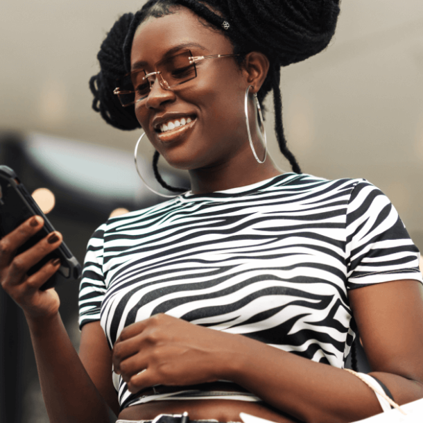 A young Black woman with space buns and large hoop earrings wears a black and white striped shirt and tinted sunglasses. She’s smiling while holding her phone in her right hand and holding shopping bags on her left arm. There are yellow and white Bokeh lights behind her.