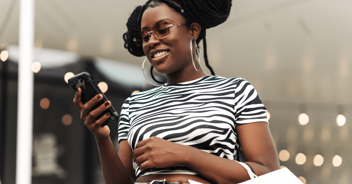 A young Black woman with space buns and large hoop earrings wears a black and white striped shirt and tinted sunglasses. She’s smiling while holding her phone in her right hand and holding shopping bags on her left arm. There are yellow and white Bokeh lights behind her.