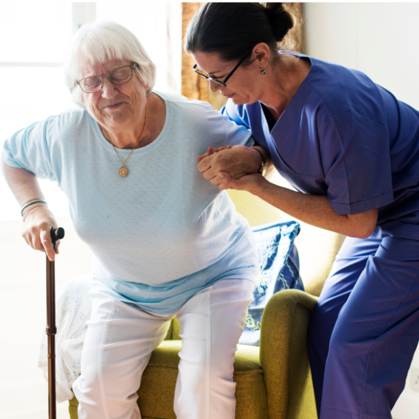 A middle-aged white woman with brown hair and blue scrubs holds hands with an old white woman with white hair and a blue shirt up out of a rocking chair. The old woman squints and strains against her can while leaning into the woman in scrubs to get up.