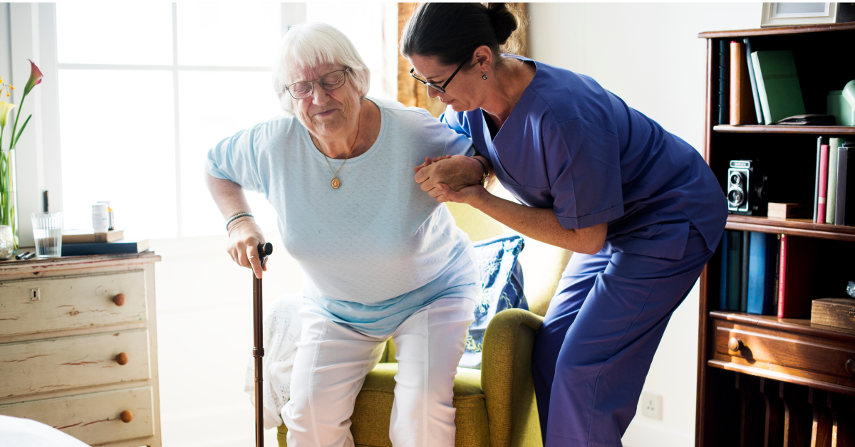 A middle-aged white woman with brown hair and blue scrubs holds hands with an old white woman with white hair and a blue shirt up out of a rocking chair. The old woman squints and strains against her can while leaning into the woman in scrubs to get up.