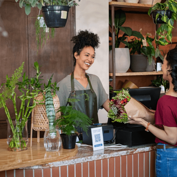 A Black woman with a top knot and kinky hair wearing a blue denim work apron smiles from behind a cash register. Across from her is a woman in a red shirt buying a bouquet of flowers. They are surrounded by green plants of all shapes and sizes.