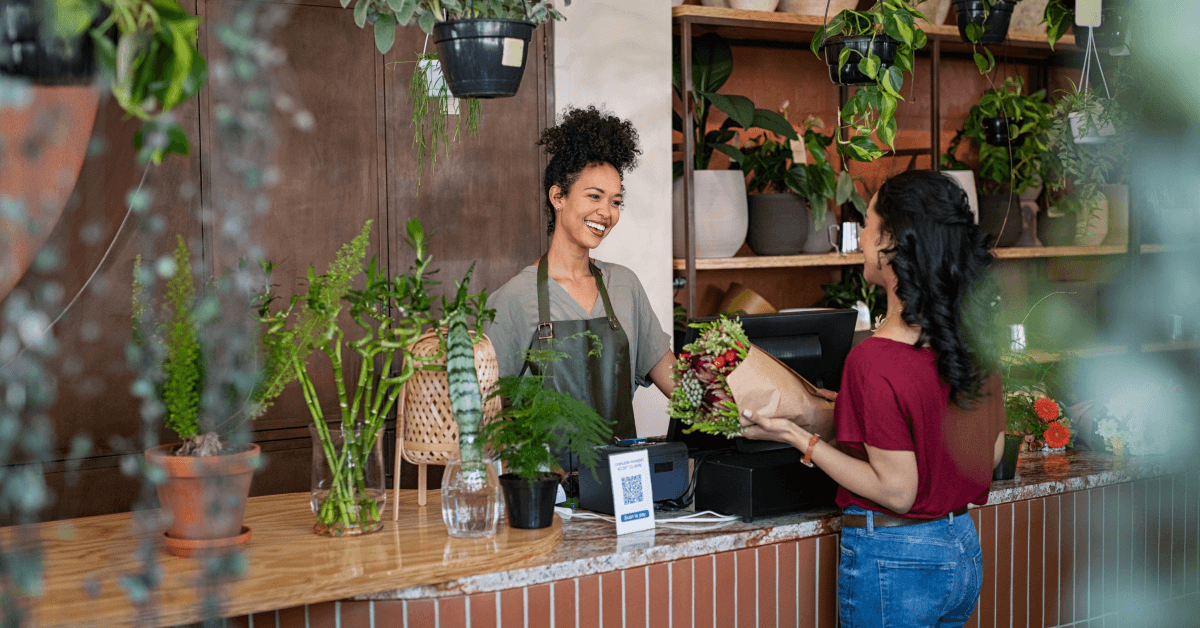 A Black woman with a top knot and kinky hair wearing a blue denim work apron smiles from behind a cash register. Across from her is a woman in a red shirt buying a bouquet of flowers. They are surrounded by green plants of all shapes and sizes.