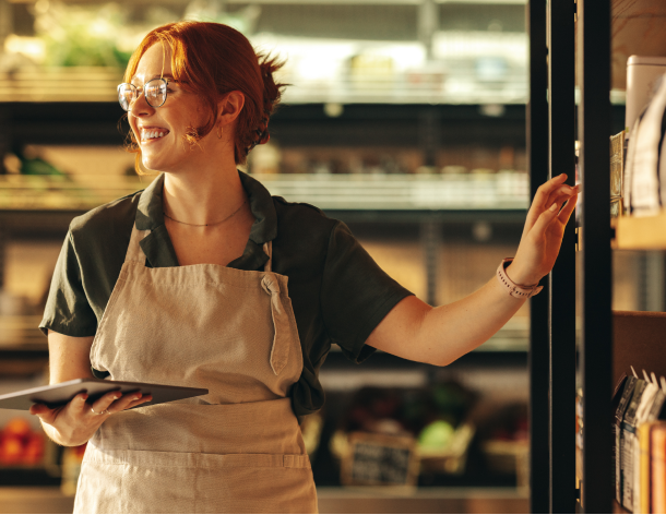 A woman wearing a work apron holding an iPad reaches for produce in a grocery store.