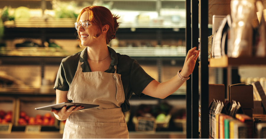 A woman wearing a work apron holding an iPad reaches for produce in a grocery store.