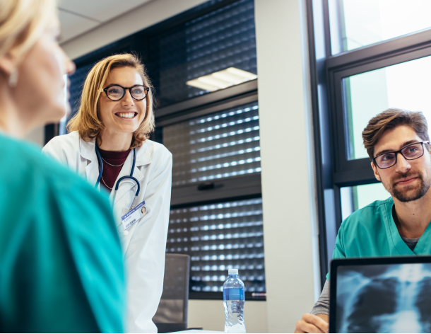 Healthcare professionals wearing scrubs sit around a conference table in a meeting. A female doctor smiles while listening to a team member speak.