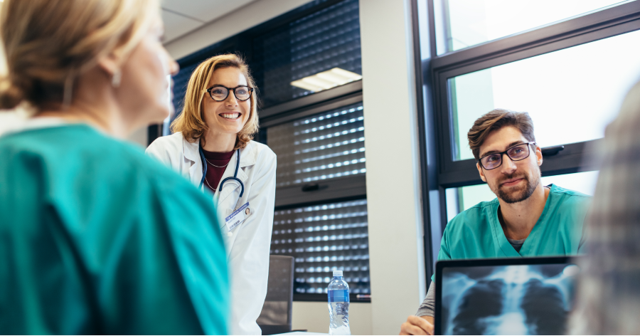 Healthcare professionals wearing scrubs sit around a conference table in a meeting. A female doctor smiles while listening to a team member speak.