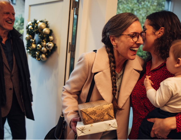 Grandmother traveling for the holidays gives daughter holding baby a one-armed hug while holding wrapped presents. Grandpa stands in the doorstop next to a Christmas wreath.