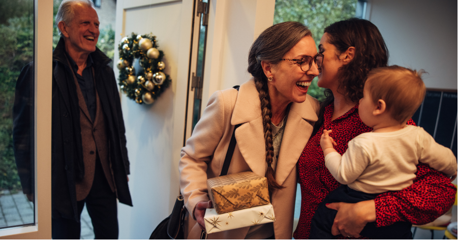 Grandmother traveling for the holidays gives daughter holding baby a one-armed hug while holding wrapped presents. Grandpa stands in the doorstop next to a Christmas wreath.