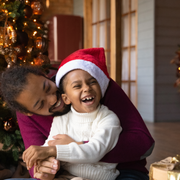 Man hugs child wearing a santa hat in front of a christmas tree.