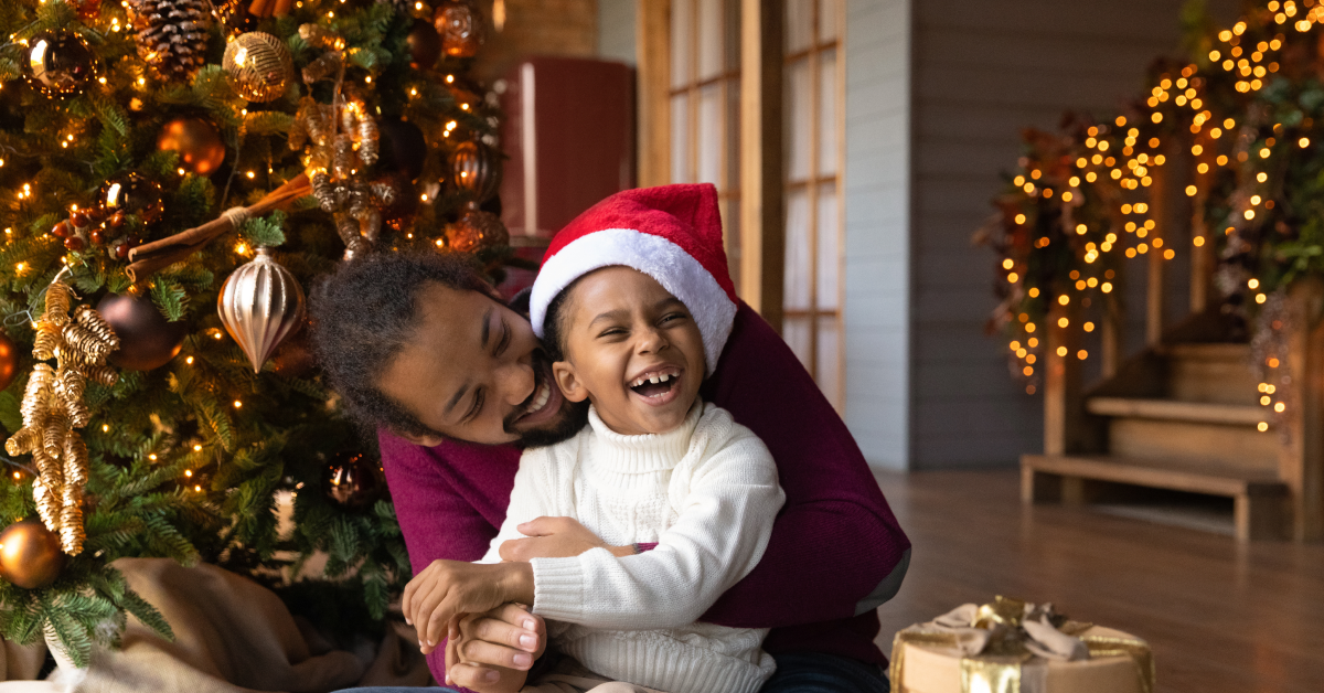 Man hugs child wearing a santa hat in front of a christmas tree.