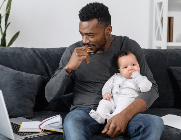 Man holding a baby while sitting on a couch thinks while enrolling in employer benefits on his computer.