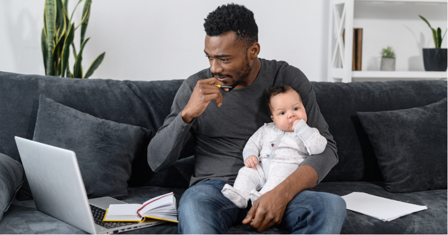 Man holding a baby while sitting on a couch thinks while enrolling in employer benefits on his computer.