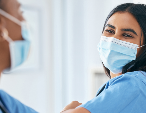 A young woman with brown hair wearing a face mask and scrubs elbow bumps another woman wearing scrubs.