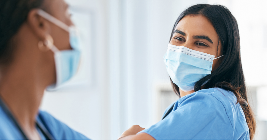 A young woman with brown hair wearing a face mask and scrubs elbow bumps another woman wearing scrubs.