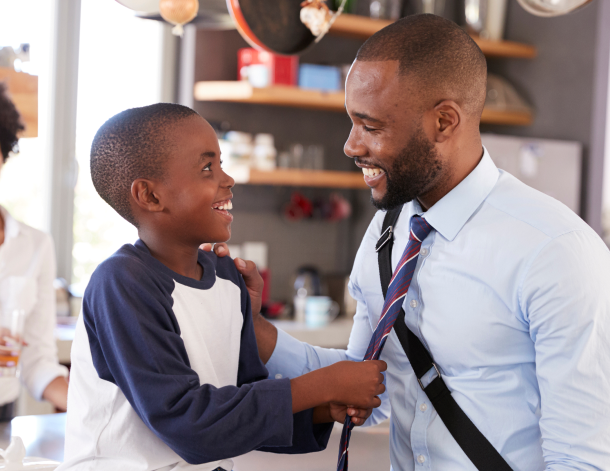 A dad on his way to a new job wears a briefcase and says goodbye to his son in the kitchen while the boy's mom smiles watching.