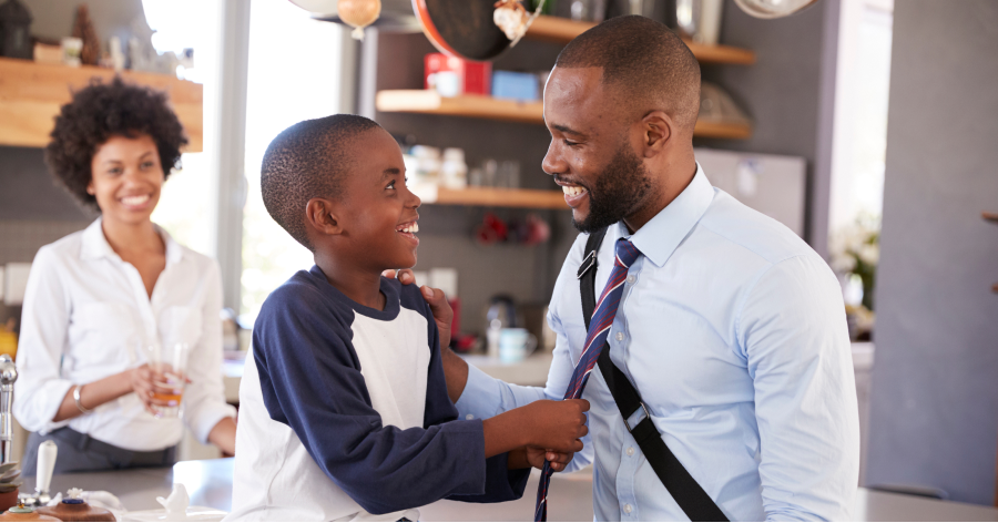 A dad on his way to a new job wears a briefcase and says goodbye to his son in the kitchen while the boy's mom smiles watching.