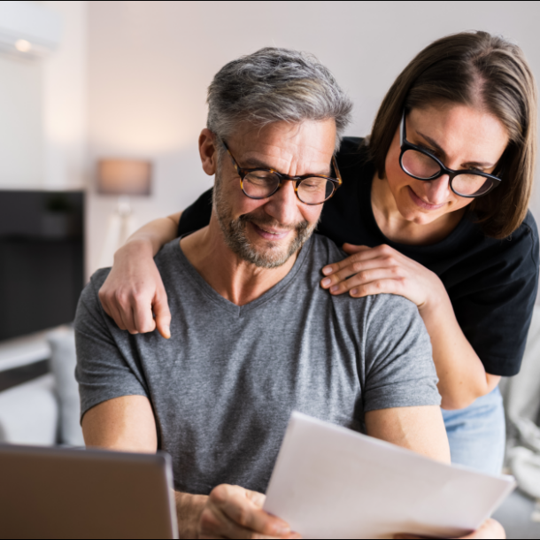 A man sits at a desk holding a piece of paper and a woman stands behind him looking. They are reading tax season tips for 2022 so they can file their taxes on-time.