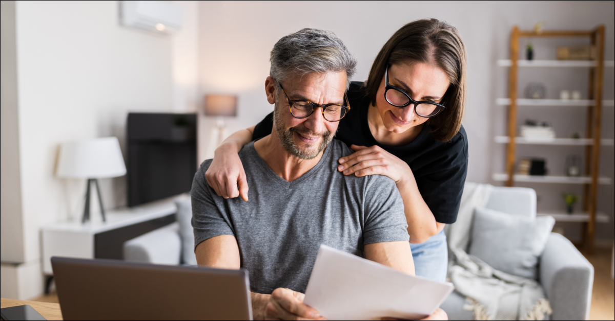 A man sits at a desk holding a piece of paper and a woman stands behind him looking. They are reading tax season tips for 2022 so they can file their taxes on-time.