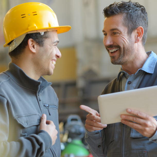 Two men working in a factory have a conversation. One is smiling and wearing a yellow hard hat and the other is holding an iPad and pointing. They are talking about financial wellness benefits.