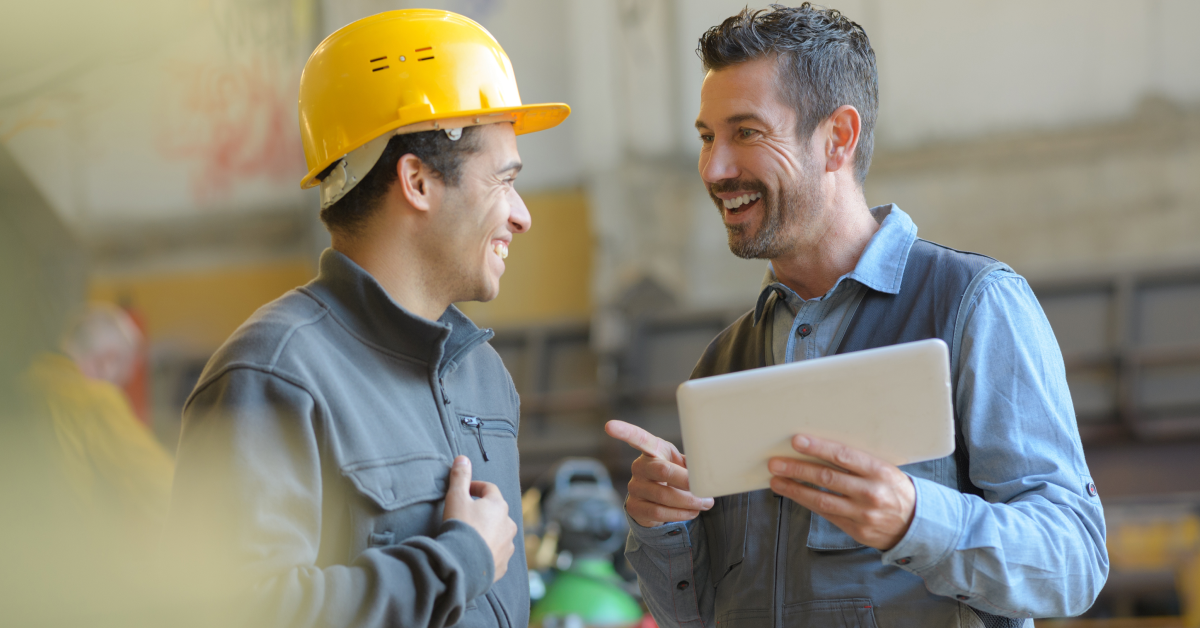 Two men working in a factory have a conversation. One is smiling and wearing a yellow hard hat and the other is holding an iPad and pointing. They are talking about financial wellness benefits.