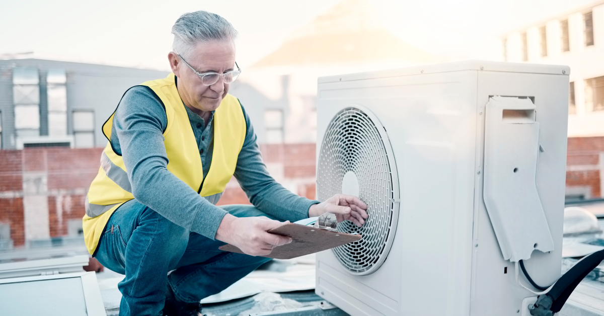 A manufacturing employee wearing a neon vest holding a clipboard monitors an HVAC system on a rooftop.