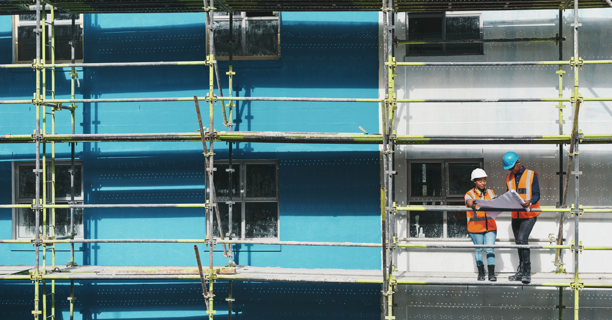 Two construction employees stand on scaffolding.