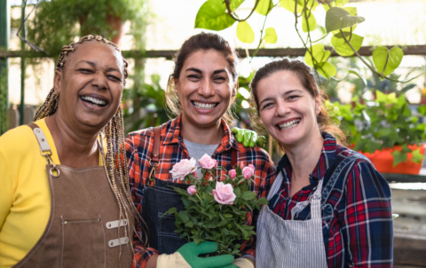 A group of three women wearing aprons pose and smile while holding flower pots in a garden nursery.