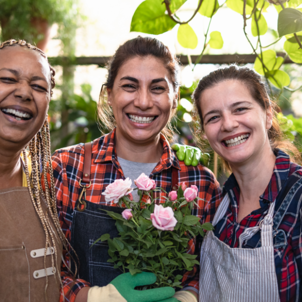 A group of three women wearing aprons pose and smile while holding flower pots in a garden nursery.