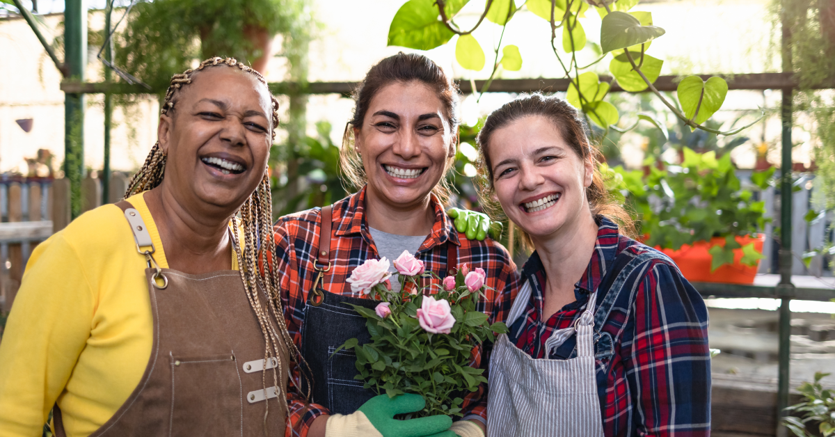 A group of three women wearing aprons pose and smile while holding flower pots in a garden nursery.
