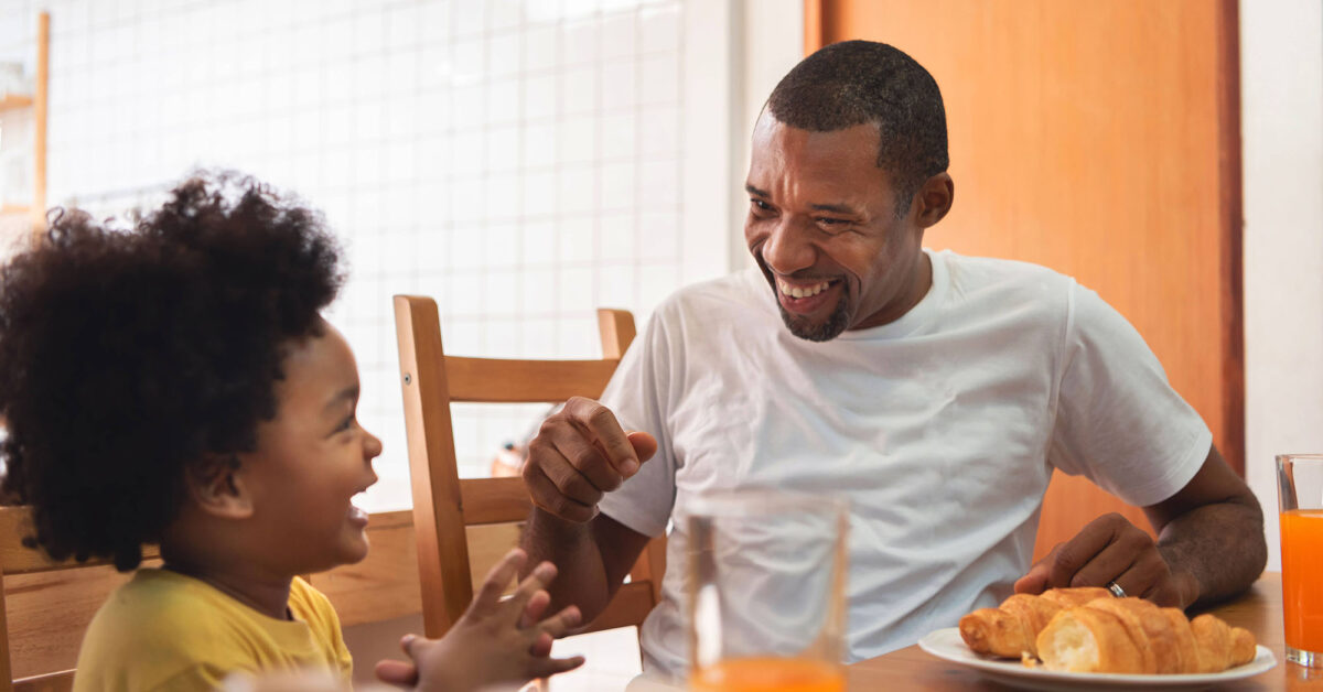 Dad and child laugh while eating breakfast.