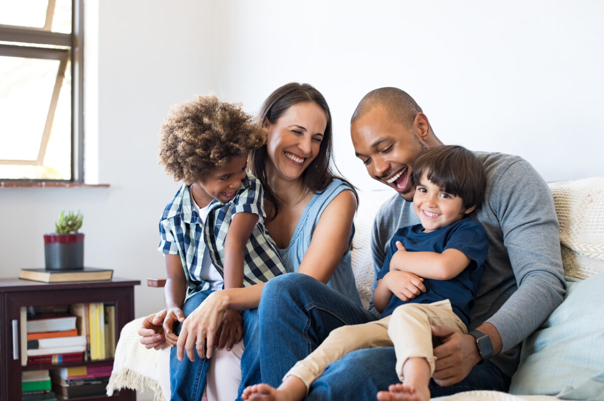 Two parents and their 2 young children sitting on the sofa and laughing with each other.