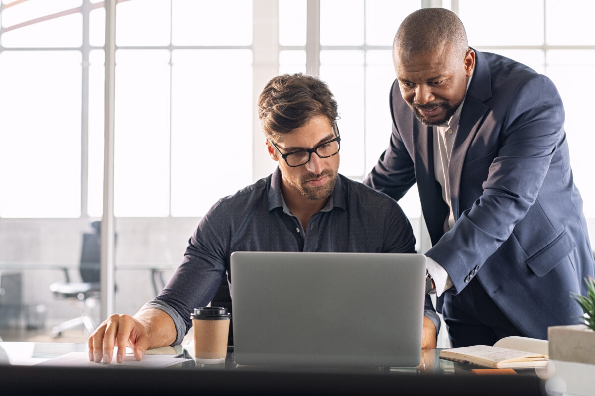 An employee is sitting with his laptop and his manager is standing next to him, helping him with his work.