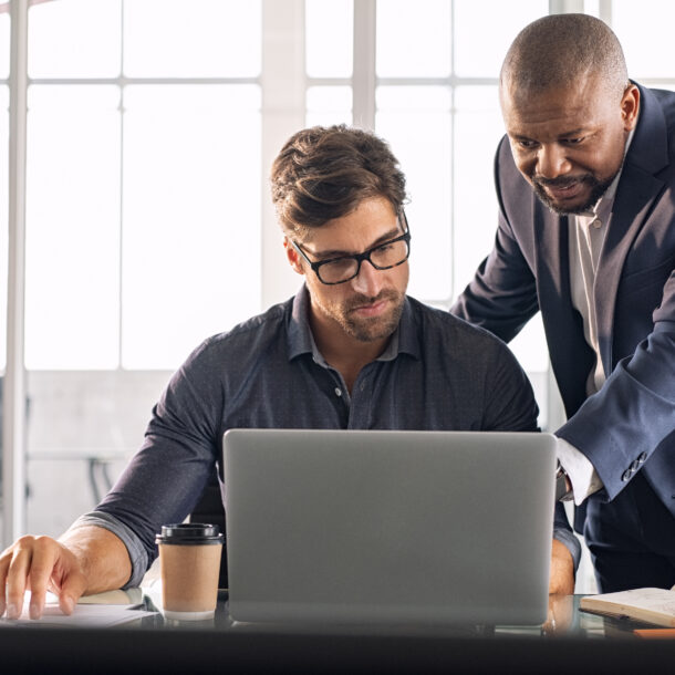 An employee is sitting with his laptop and his manager is standing next to him, helping him with his work.