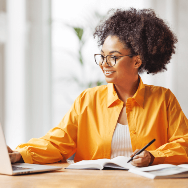 A woman with glasses is at a desk with her laptop and a notebook.