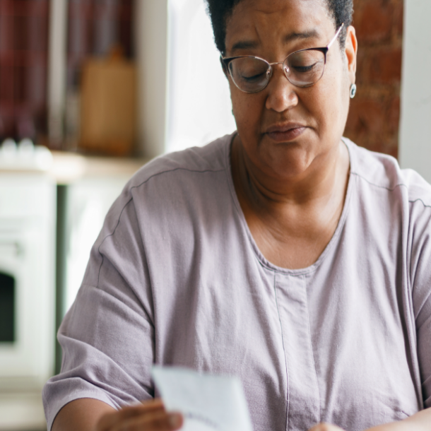 Portrait of puzzled grandma in glasses reading paycheck or utility bill attentively sitting in her kitchen in front of laptop and smartphone