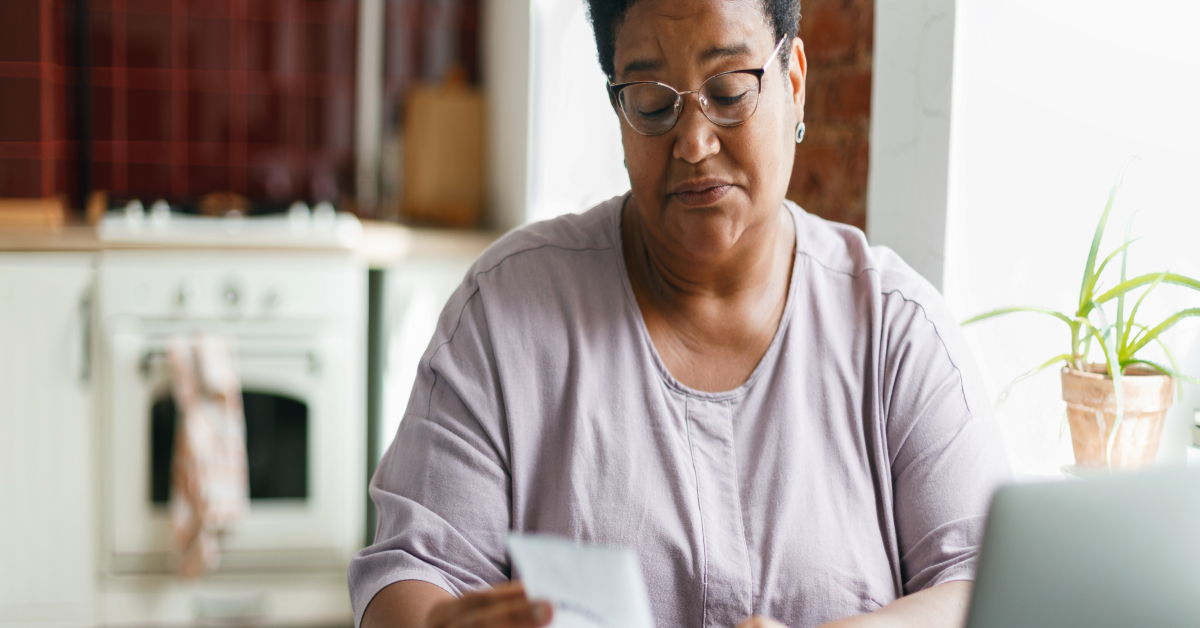 Portrait of puzzled grandma in glasses reading paycheck or utility bill attentively sitting in her kitchen in front of laptop and smartphone