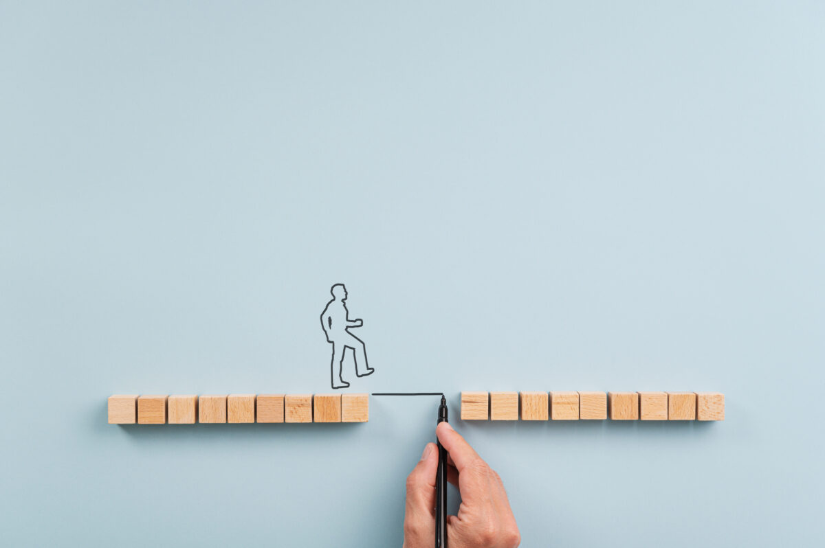 Male hand drawing a bridge between two rows of wooden blocks for a silhouetted man to walk across. Over blue background with plenty of copy space.